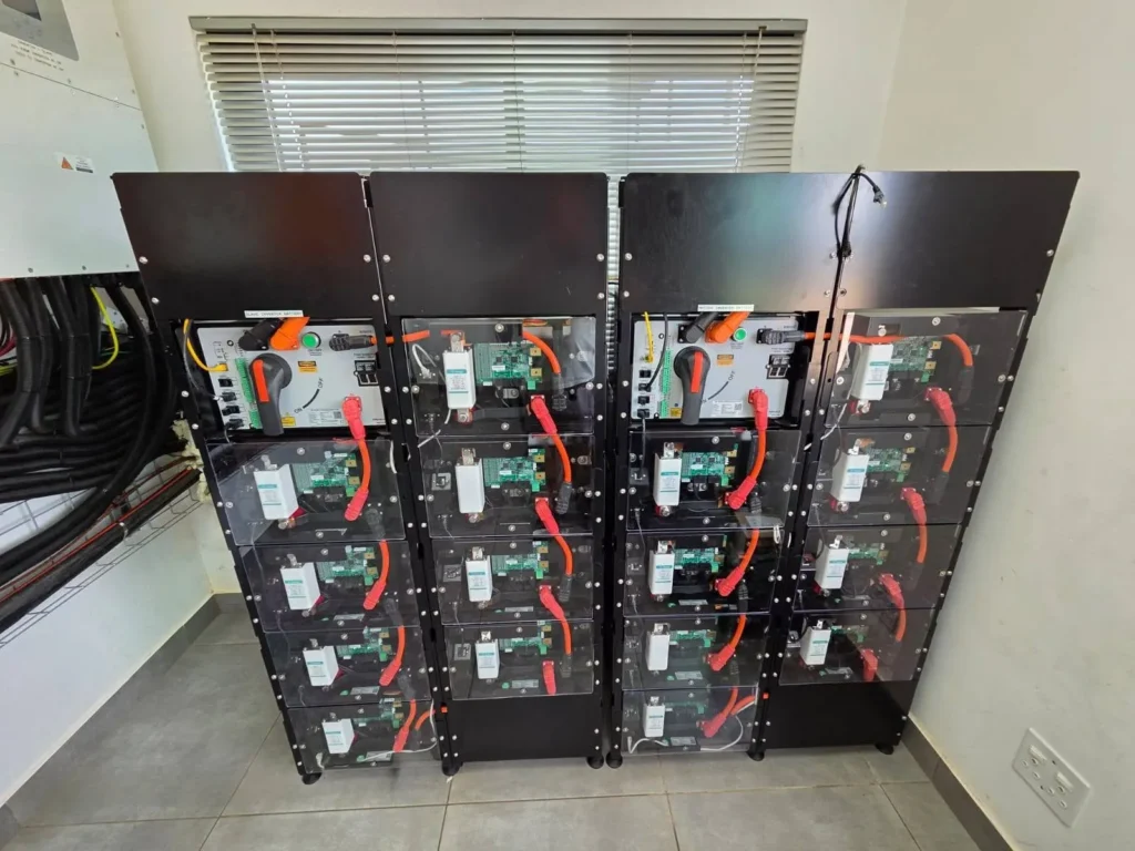 Four stacks of modular battery units side by side in a white room, each with an isolator switch and orange power cables