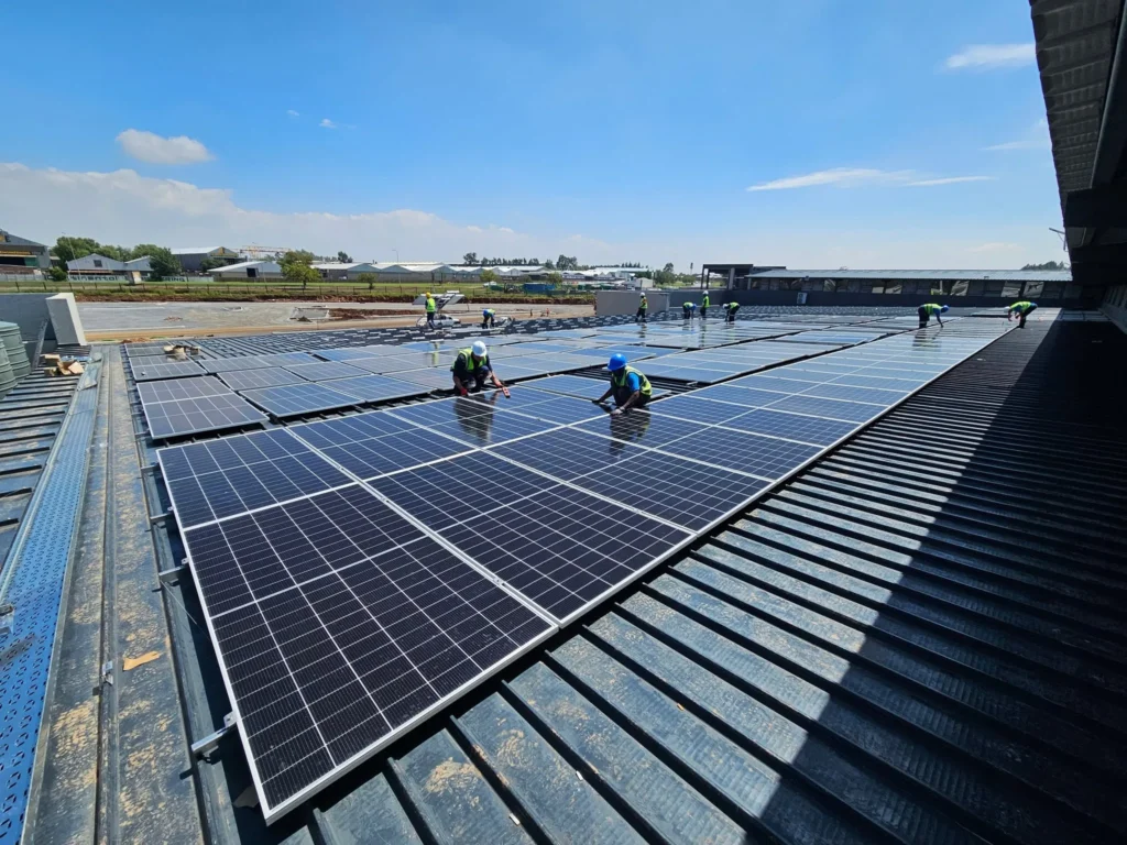 Installation crew in hi-vis vests kneeling on a commercial roof to fix solar panels in long rows under a blue sky