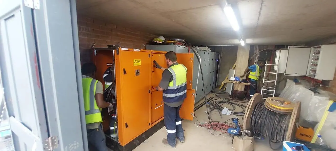 Electricians in hi-vis vests working at an open orange switchgear panel in a plant room with a cable drum on the floor