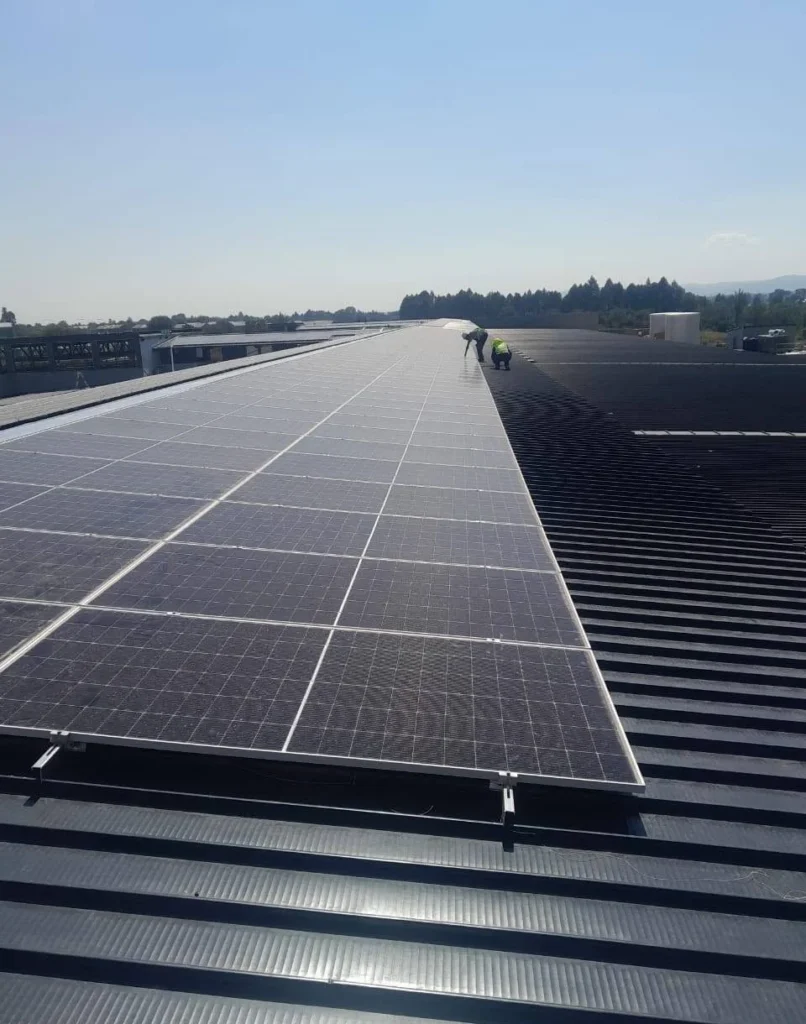 Long rows of solar panels on a dark profiled metal roof with two installers working at the far end