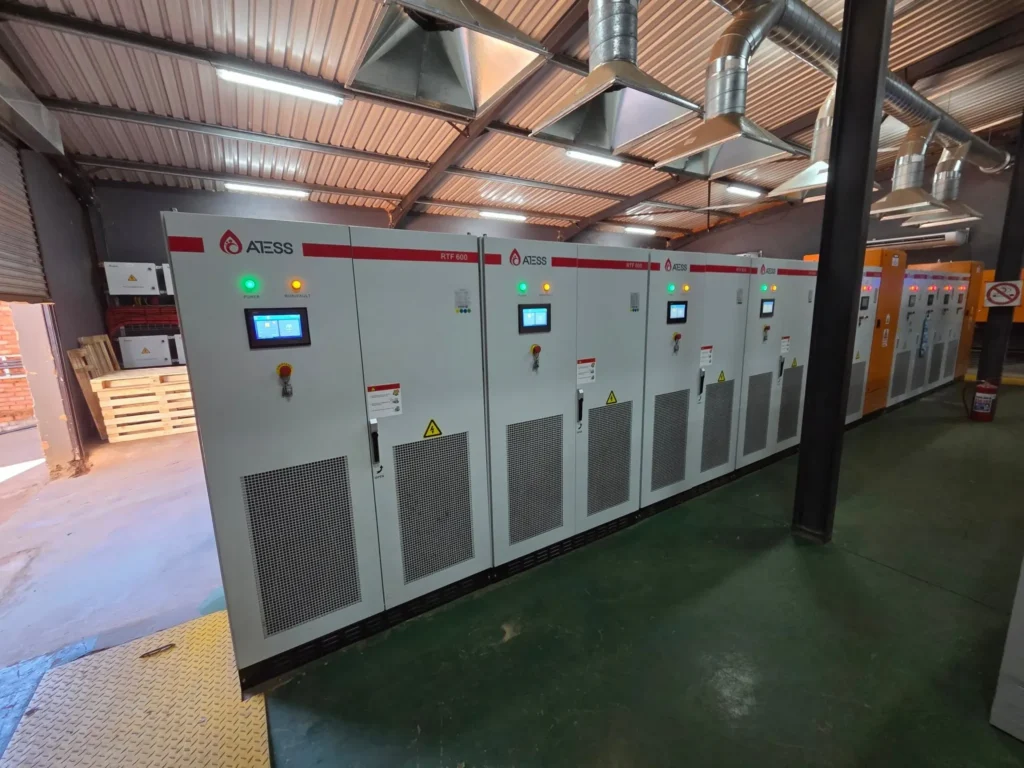 Row of white ATESS power conversion cabinets with touchscreens and status lights beside orange switchgear on a green floor