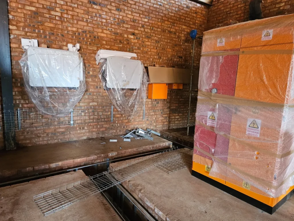 Two solar inverters and orange DC boxes on a brick wall, still in protective plastic, with a wrapped switchboard beside them